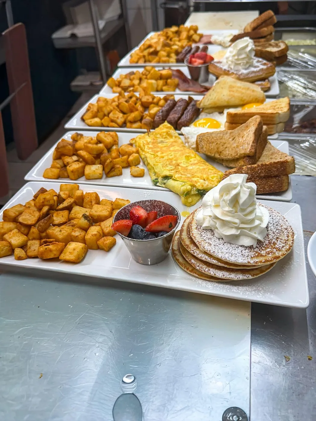 Breakfast plates ready on the pass — home fries, omelette, toast, pancakes and fresh fruit.